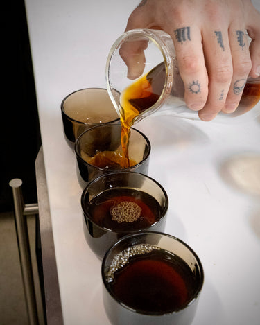 Hand pouring coffee into a series of small glass cups on a white surface.
