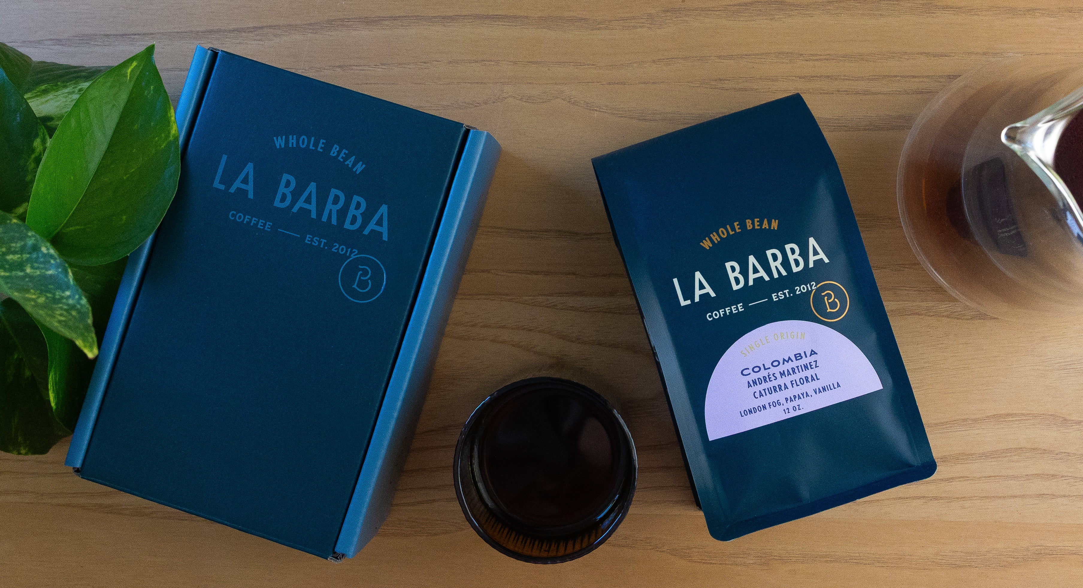 Two blue coffee packages labeled 'La Barba' on a wooden surface with a plant and glass of water in the background.