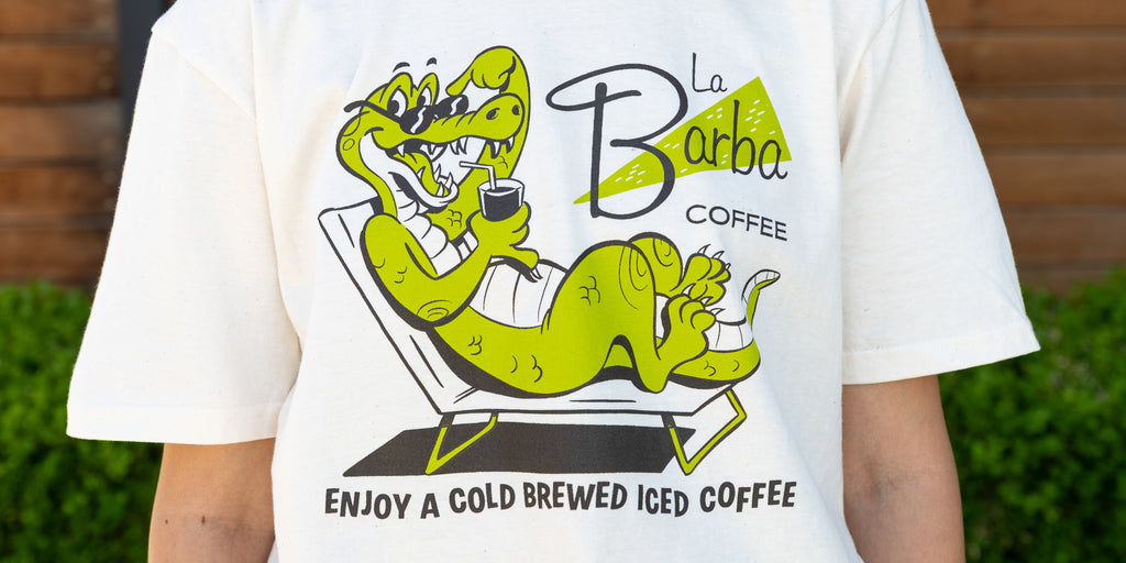 White t-shirt with a green alligator graphic and 'La Barba Coffee' text, worn by a person outdoors.