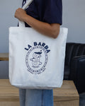 Person holding a white tote bag with 'La Barba' logo