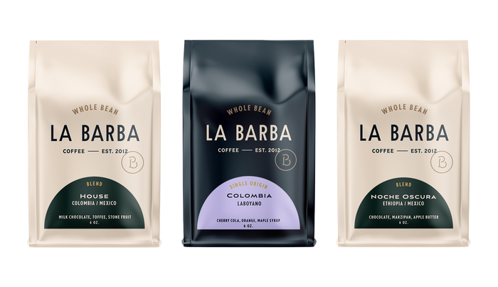 A product photo featuring three La Barba Coffee bags displayed side by side. The left bag is the La Barba House Blend (Colombia/Mexico) in a light beige package with tasting notes of milk chocolate, toffee, and stone fruit. The center bag is a dark navy single origin Colombia Laboyano coffee with tasting notes of cherry cola, orange, and maple syrup. The right bag is the Noche Oscura Blend (Ethiopia/Mexico) in a matching beige package with notes of chocolate, marzipan, and apple butter. All bags are whole-b