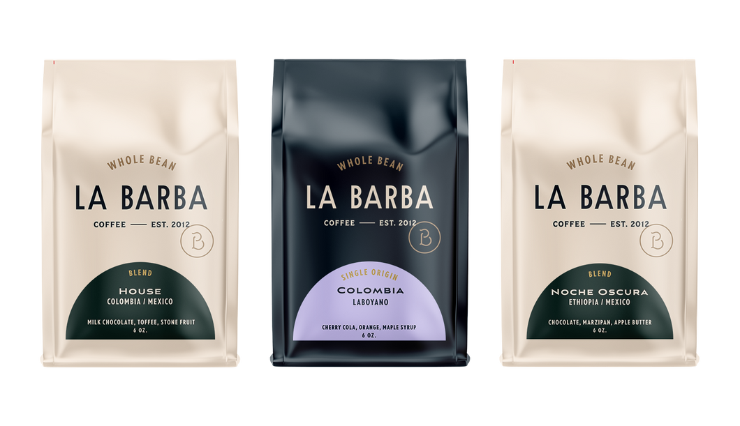 A product photo featuring three La Barba Coffee bags displayed side by side. The left bag is the La Barba House Blend (Colombia/Mexico) in a light beige package with tasting notes of milk chocolate, toffee, and stone fruit. The center bag is a dark navy single origin Colombia Laboyano coffee with tasting notes of cherry cola, orange, and maple syrup. The right bag is the Noche Oscura Blend (Ethiopia/Mexico) in a matching beige package with notes of chocolate, marzipan, and apple butter. All bags are whole-b