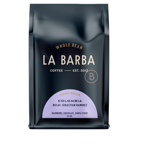 The image features a 12oz bag of La Barba Coffee, specifically their Colombia Decaf Sebastian Ramirez. The bag is designed with a dark background, prominently displaying the brand name "La Barba" in large, elegant letters. It highlights the coffee's origin, Colombia, along with a description of the flavor profile, including notes of orange zest, chocolate, and nougat. The design exudes a premium and modern feel with a minimalist, sophisticated look.