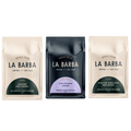 A product photo featuring three La Barba Coffee bags displayed side by side. The left bag is the La Barba House Blend (Peru/Colombia) in a light beige package with tasting notes of milk chocolate, toffee, and stone fruit. The center bag is a dark navy single origin Colombia Laboyano coffee with tasting notes of cherry cola, orange, and maple syrup. The right bag is the Noche Oscura Blend (Peru/Ethiopia) in a matching beige package with notes of chocolate, marzipan, and apple butter.